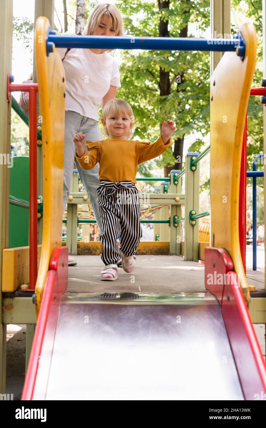 A beautiful cheerful little girl rides a childrens city slide in park. Mom helps not to fall. Lifestyle Stock Photo