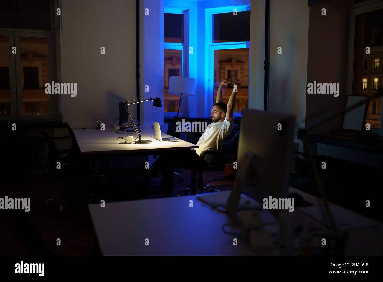 Remote programmer work late night stretching hands while sitting at laptop computer in dark office Stock Photo