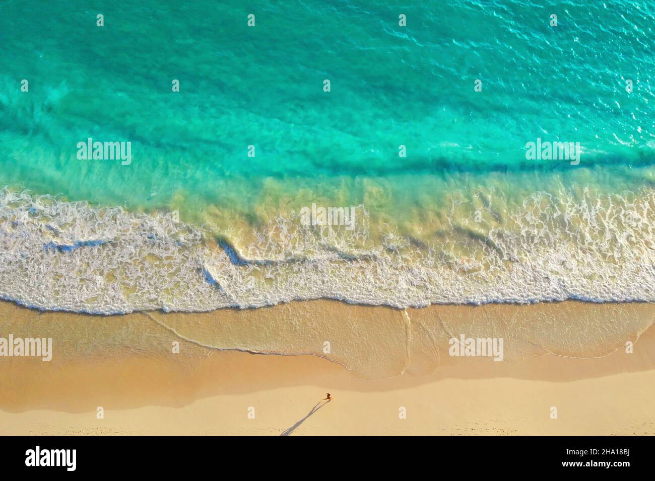 Man running along the ocean coast, top view. Overhead view of man run by the white sandy beach along the amazing clear light blue ocean. Run along the coast of the ocean at sunrise. Healthy lifestyle. Stock Photo