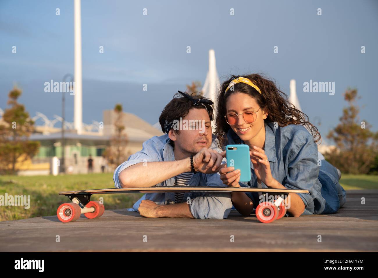 Happy smiling woman show man message on smartphone lying on skateboard outdoor in summer urban park Stock Photo