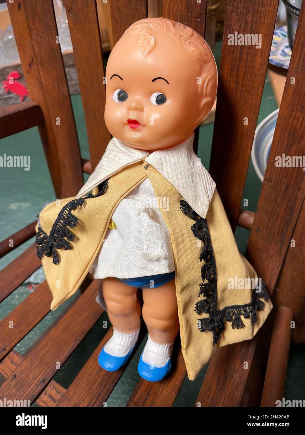 A vintage molded plastic baby doll wearing blue shoes, a tan cape with lace trim, and an adorable suspicious expression on her face stands in a chair. Stock Photo