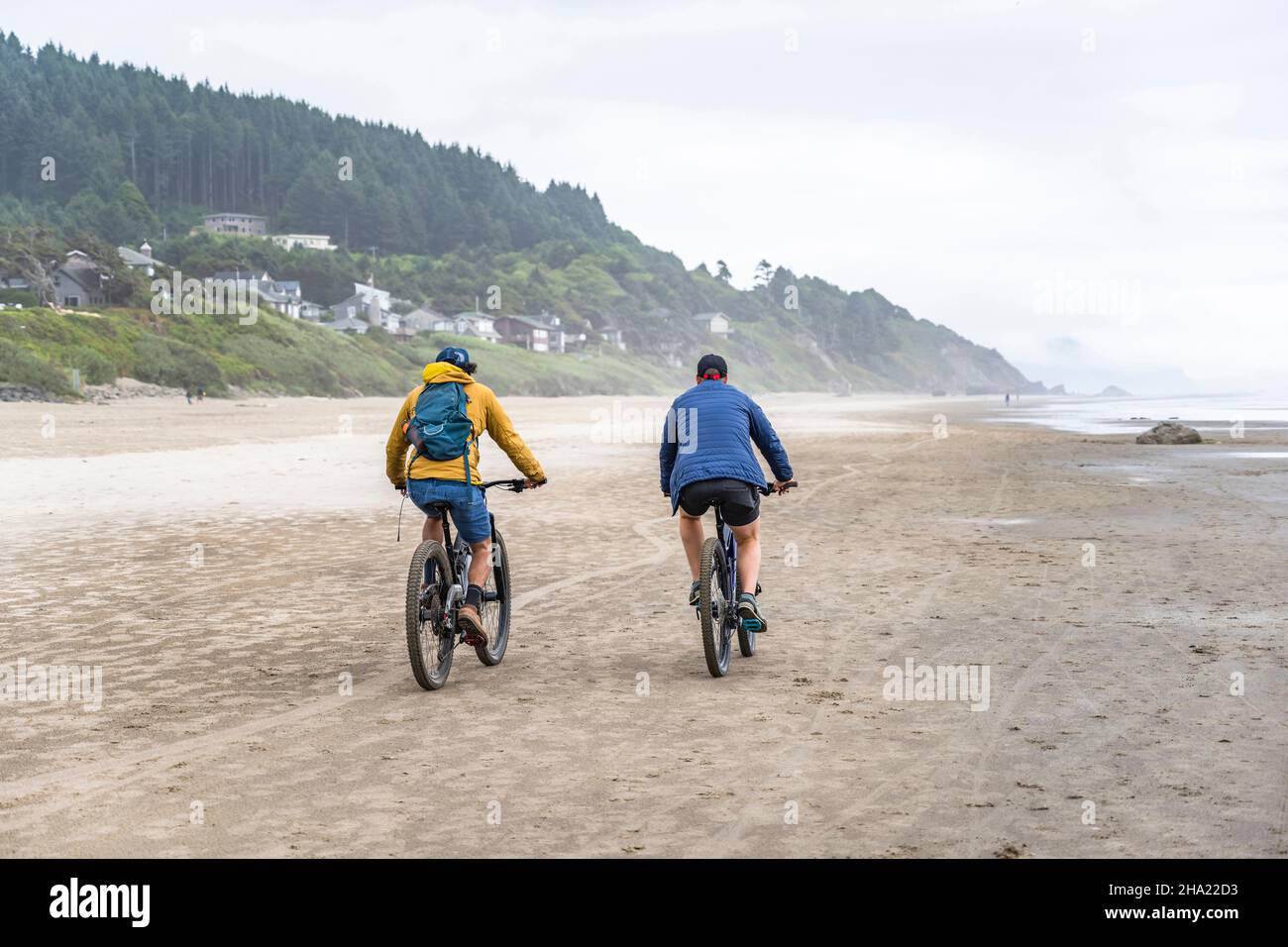 Two amateur men in in warm clothes travel side by side on the mountain bicycles along the foggy Northwest Pacific Ocean preferring an active healthy l Stock Photo