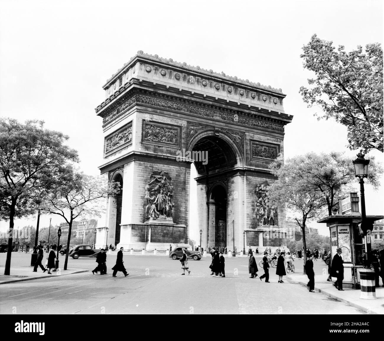 Paris Arc de triomphe 1945 with pedestrians, cylists, and few autos The view of the arch and the circular Place de l'Étoile is taken from Avenue Marceau near the end of World War II in Europe.  The roadway contains only several motor vehicles and several bicyclists.  In the photo are numerous pedestrians in the foreground and visitors beneath the Arc. There are two kiosks promoting the Loterie Nationale. Vintage streetlights are in view. The photo is framed by trees with Spring foliage. Stock Photo