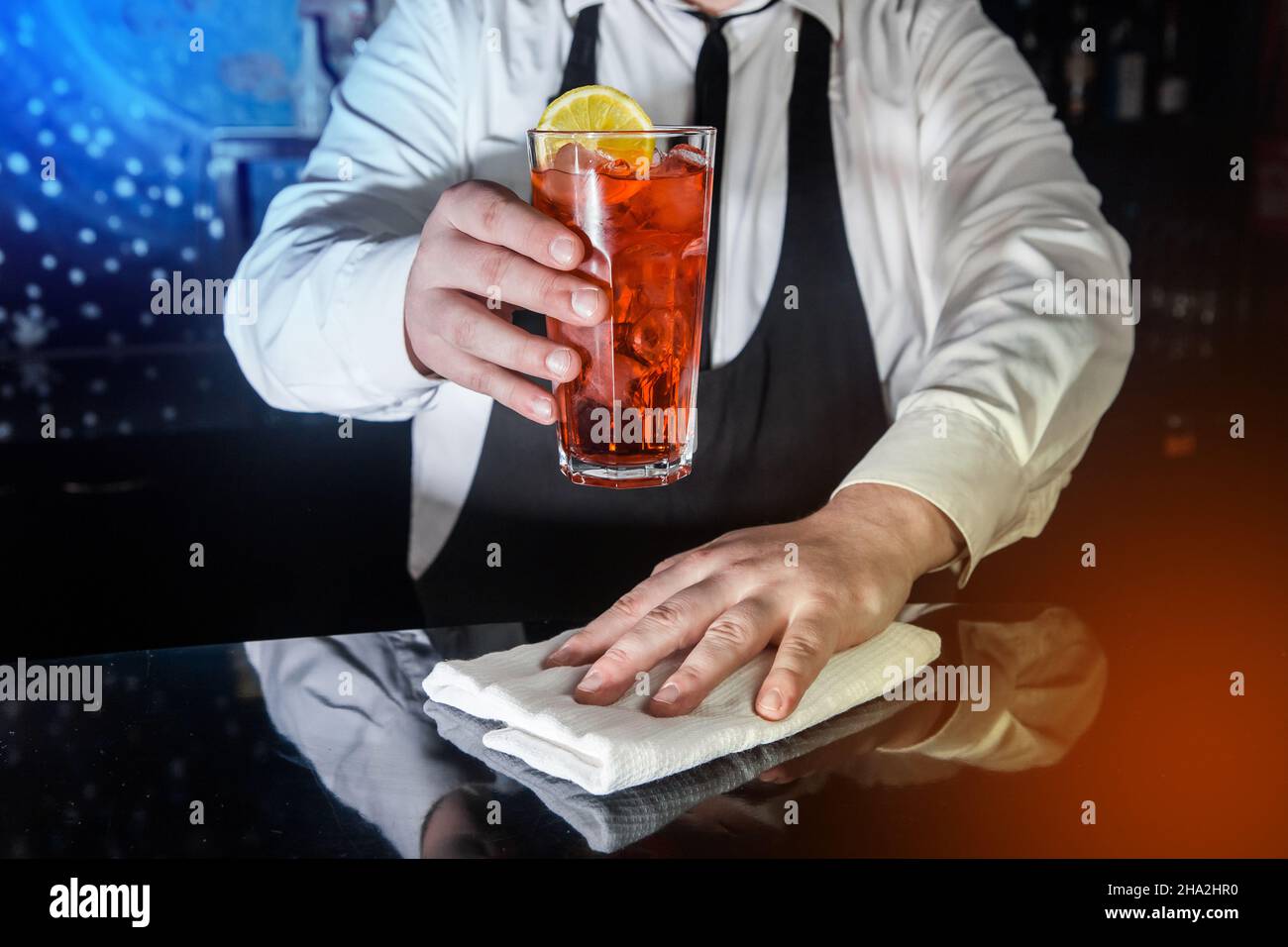 A professional bartender holds in his hand a red alcoholic cocktail, chilled drink in glass decorated with a slice of sliced lemon and wipes the bar c Stock Photo