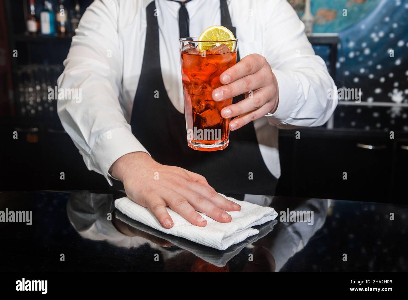 A professional bartender holds in his hand a red alcoholic cocktail, chilled drink in glass decorated with a slice of sliced lemon and wipes the bar c Stock Photo