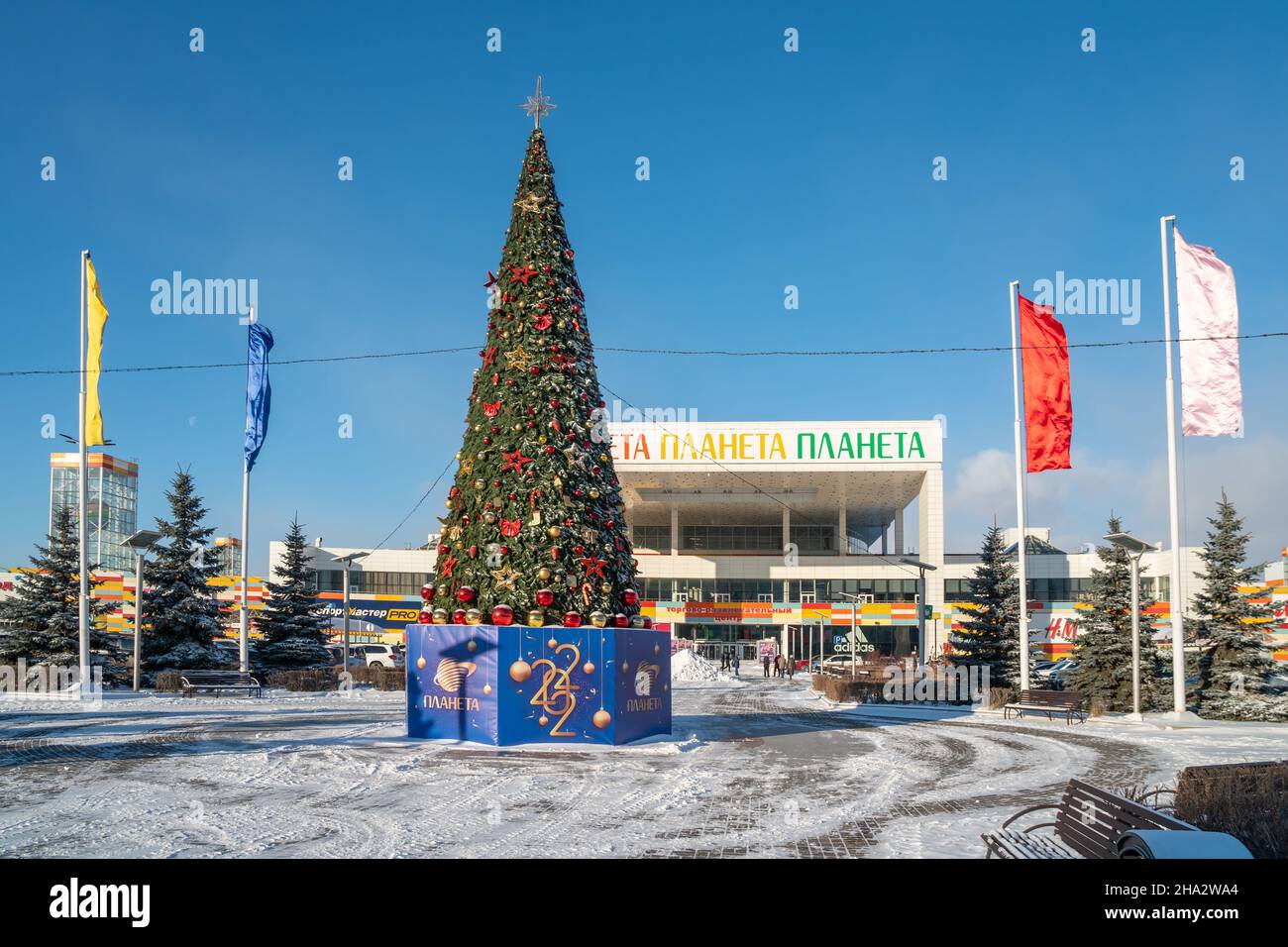 A decorated Christmas tree with the inscription Planet in Russian stands in front of the Planet shopping and entertainment center on a sunny day. Stock Photo