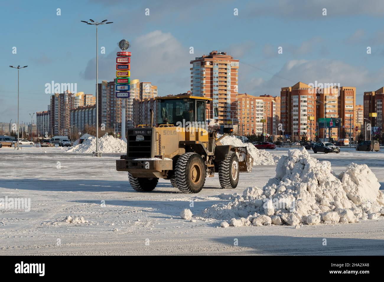 A bucket loader removes snow from snowdrifts in a city square after a snowfall against the backdrop of residential buildings on a sunny day. Stock Photo
