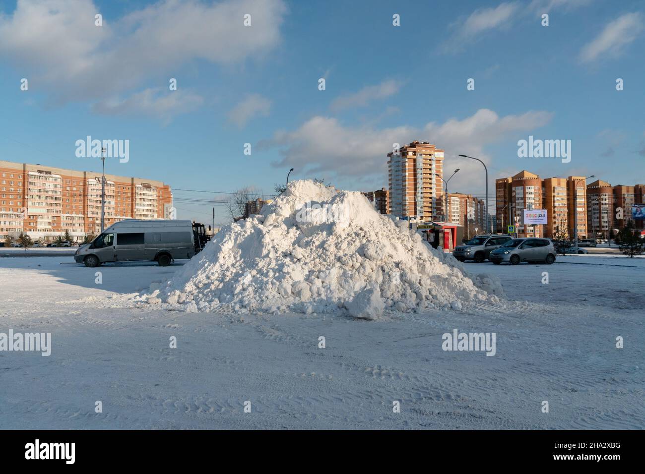 A large pile of snow lies in a parking lot on the street after a snowfall with residential buildings in the background. Stock Photo