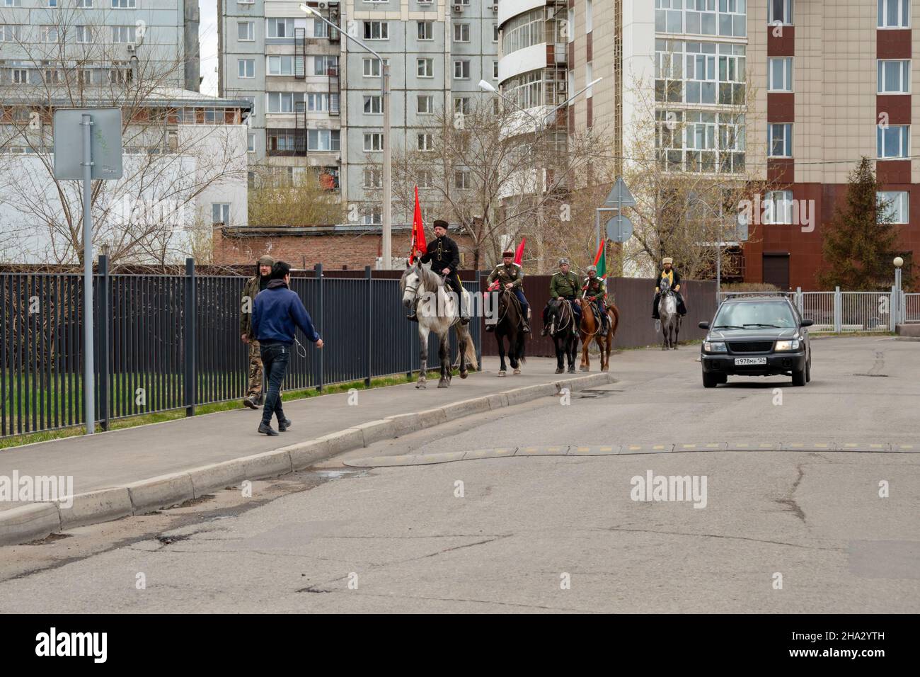 A group of Cossacks with flags rides on horseback along the sidewalk against the backdrop of residential buildings in the spring city. Stock Photo