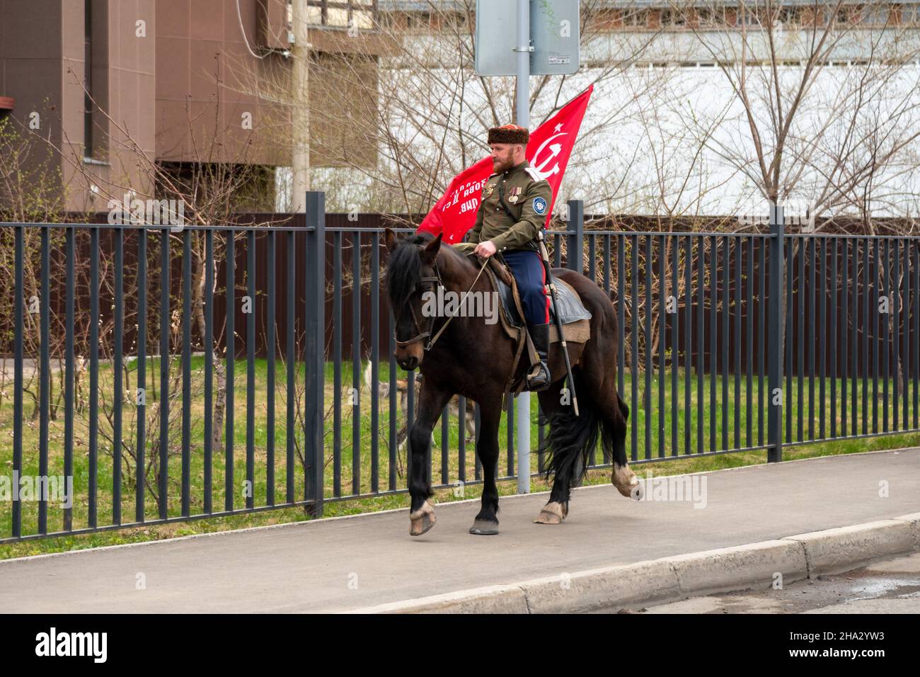 A Cossack with the regiment's banner rides a brown horse along the sidewalk along a metal fence in a spring city. Stock Photo