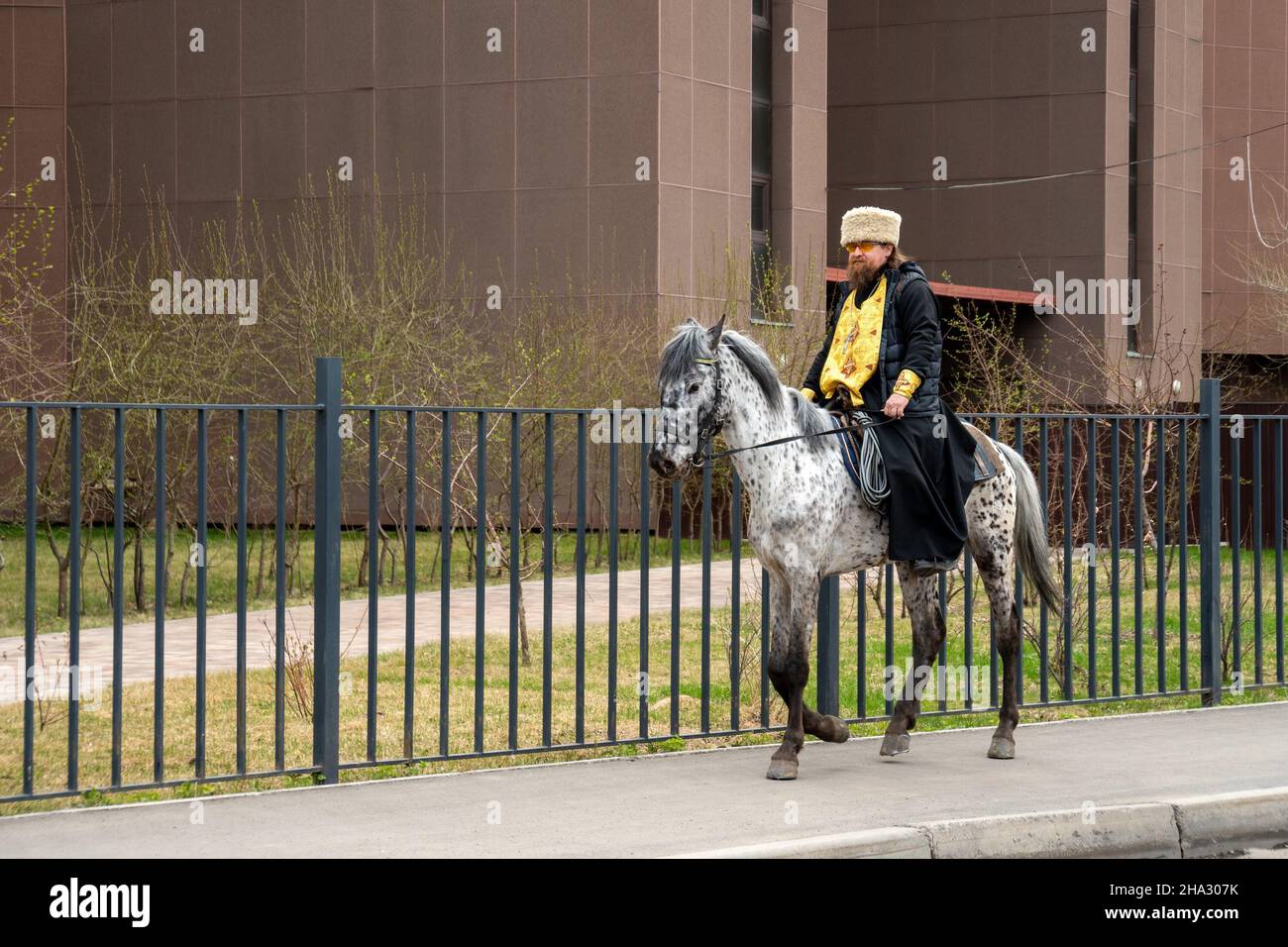 A clergyman in a cassock and hat, with a cross on his chest, rides a horse along the sidewalk along a metal fence in the spring. Stock Photo