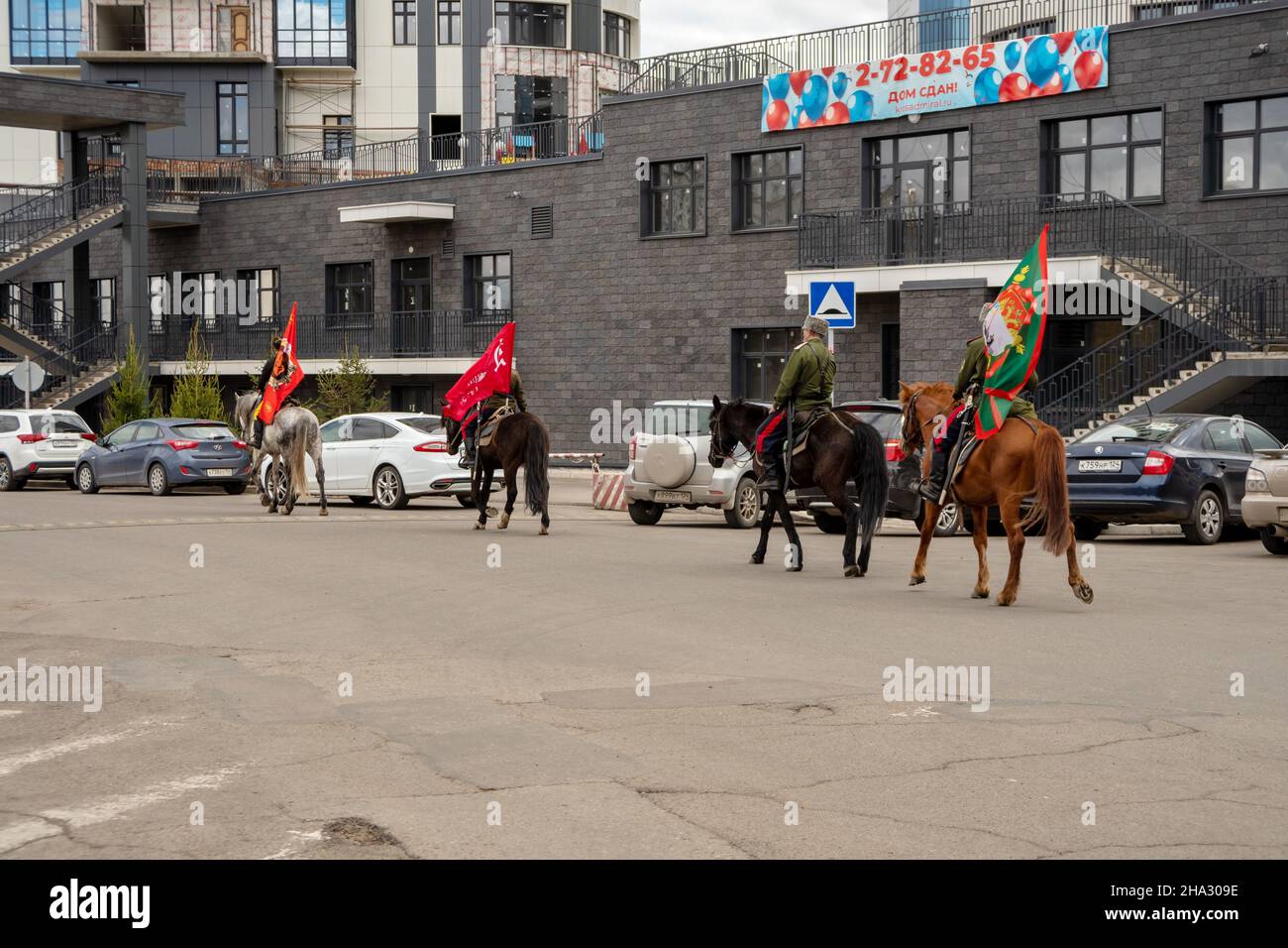 Four Cossacks with flags in their hands ride horses past parked cars in the courtyard of a residential building in the spring city. Stock Photo