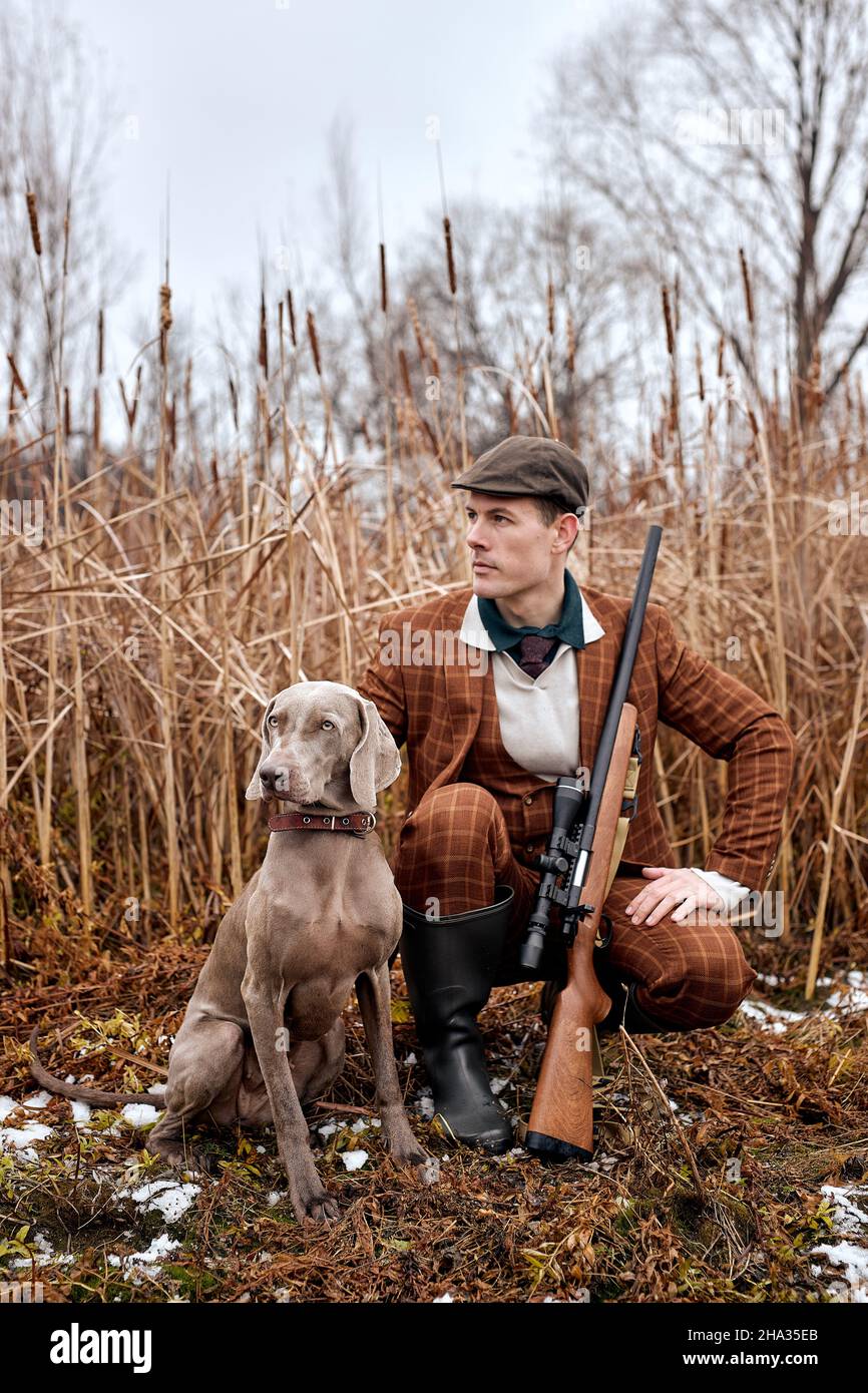 Confident american hunter man with dog holding shotgun rifle walking in countryside nature, wearing brown trendy stylish suit and hat. professional sk Stock Photo