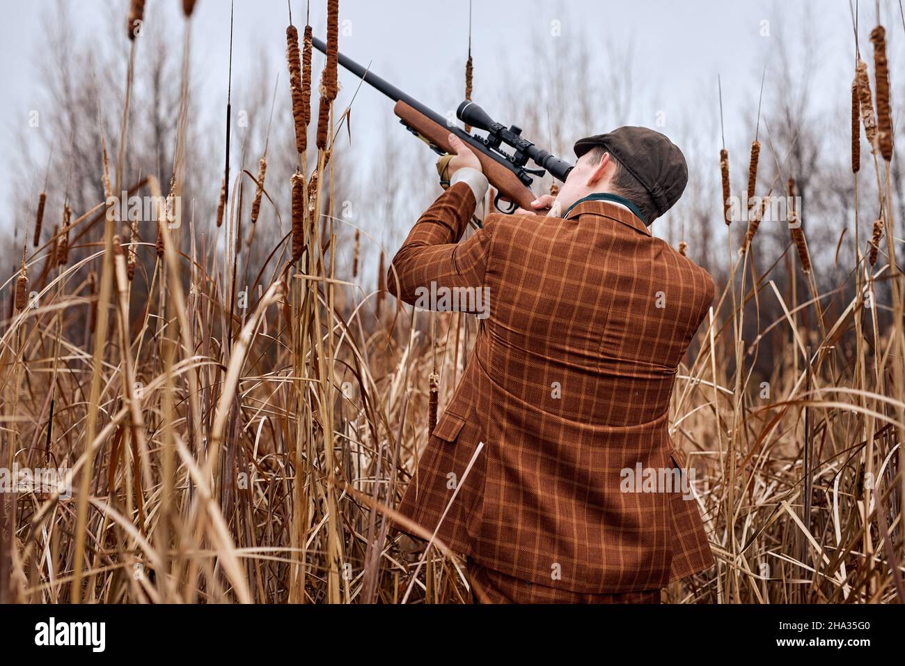 european handsome hunter is hunting in forest, looking through the rifle, soft focus. in countryside. male in trendy brown suit confidently aiming rif Stock Photo