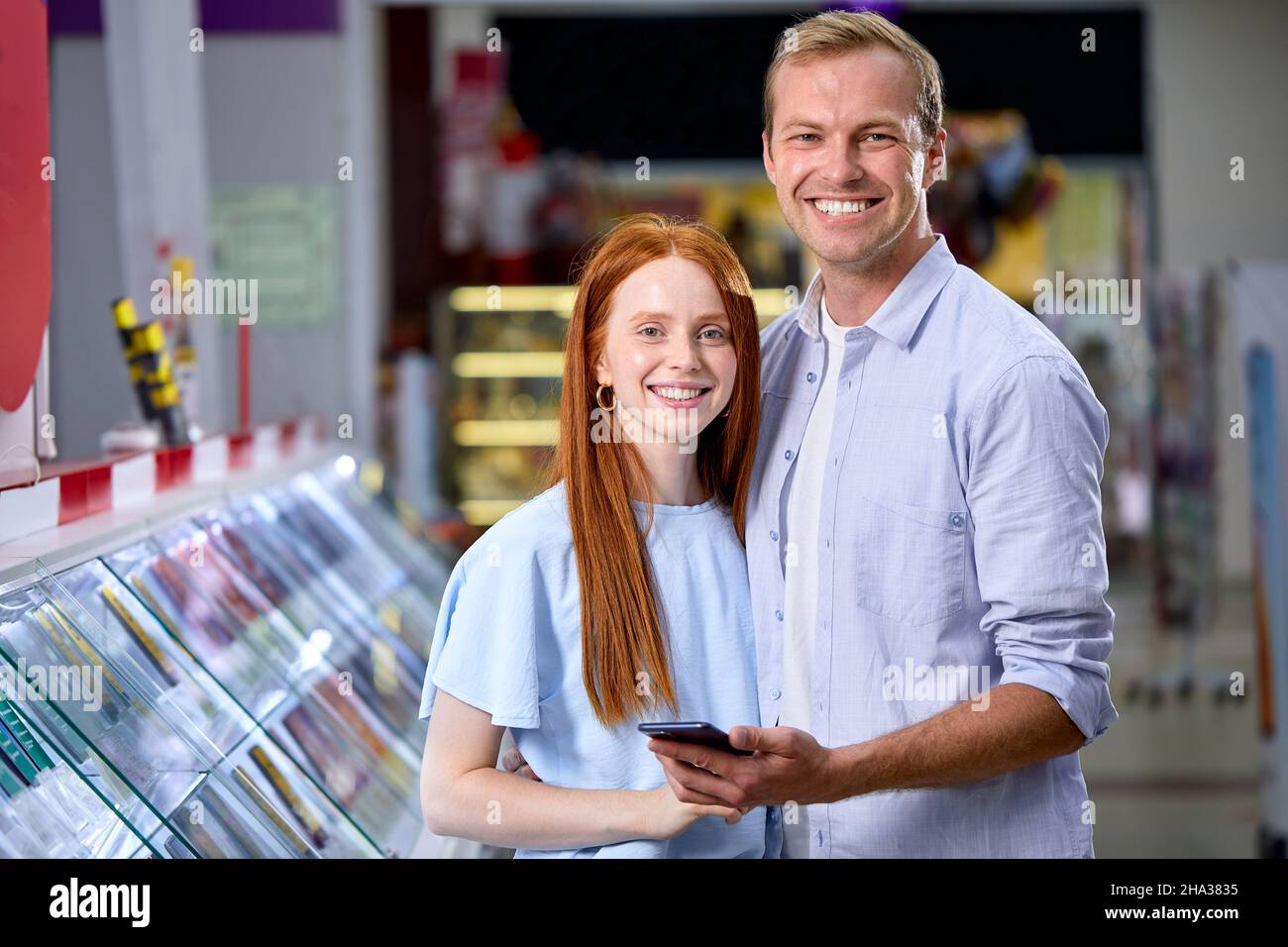 cheerful young man and woman checking out mobile phone in tech store, make choice, want to buy, consumerism concept. portrait of caucasian couple stan Stock Photo