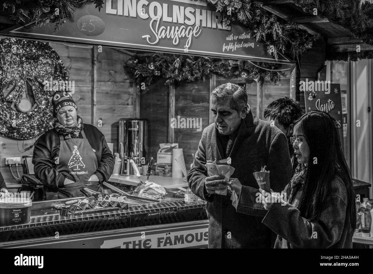 Harrogate couple eating Lincolnshire sausages and other fresh food at a market stall as the vendor watches on, North Yorkshire, England, UK. Stock Photo