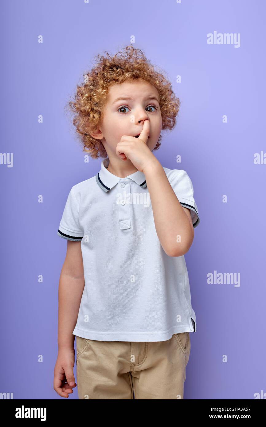 cute little curly boy picking his nose to explore it and act mischievous for fun childhood and bad habit, isolated on purple studio background, portra Stock Photo