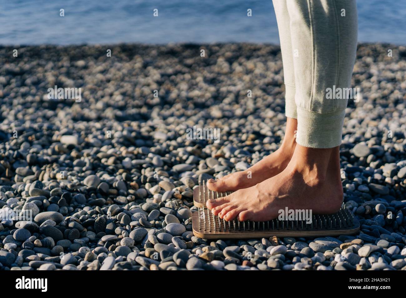 Alternative medicine acupressure, barefoot woman stand on wooden planks with nails on pebbles on the beach. Stock Photo