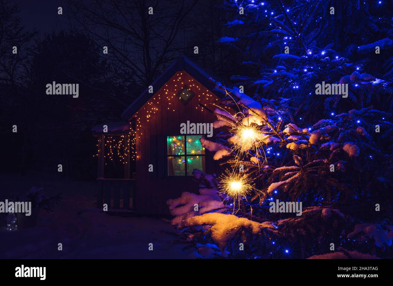 Selective focus on sparkler candles hanging and burning on spruce tree outdoors on snowy winter night. Cute decorated playhouse on background. Stock Photo