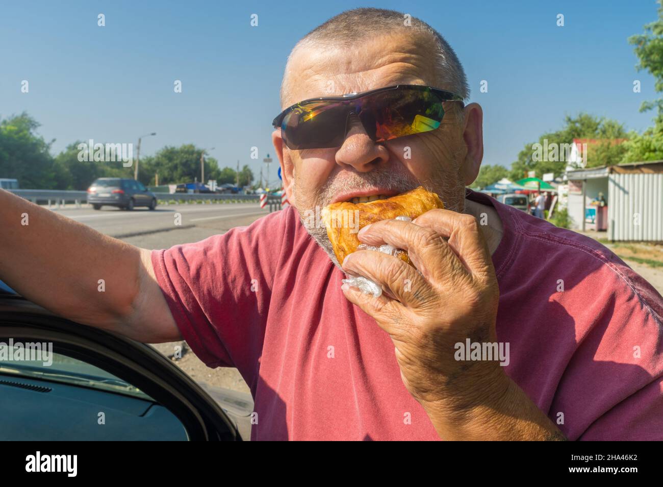 Portarait of hungry Caucasian senior driver in sunglasses eating junk food  (fried patty) near his car while doing short stop on Ukrainian road Stock Photo