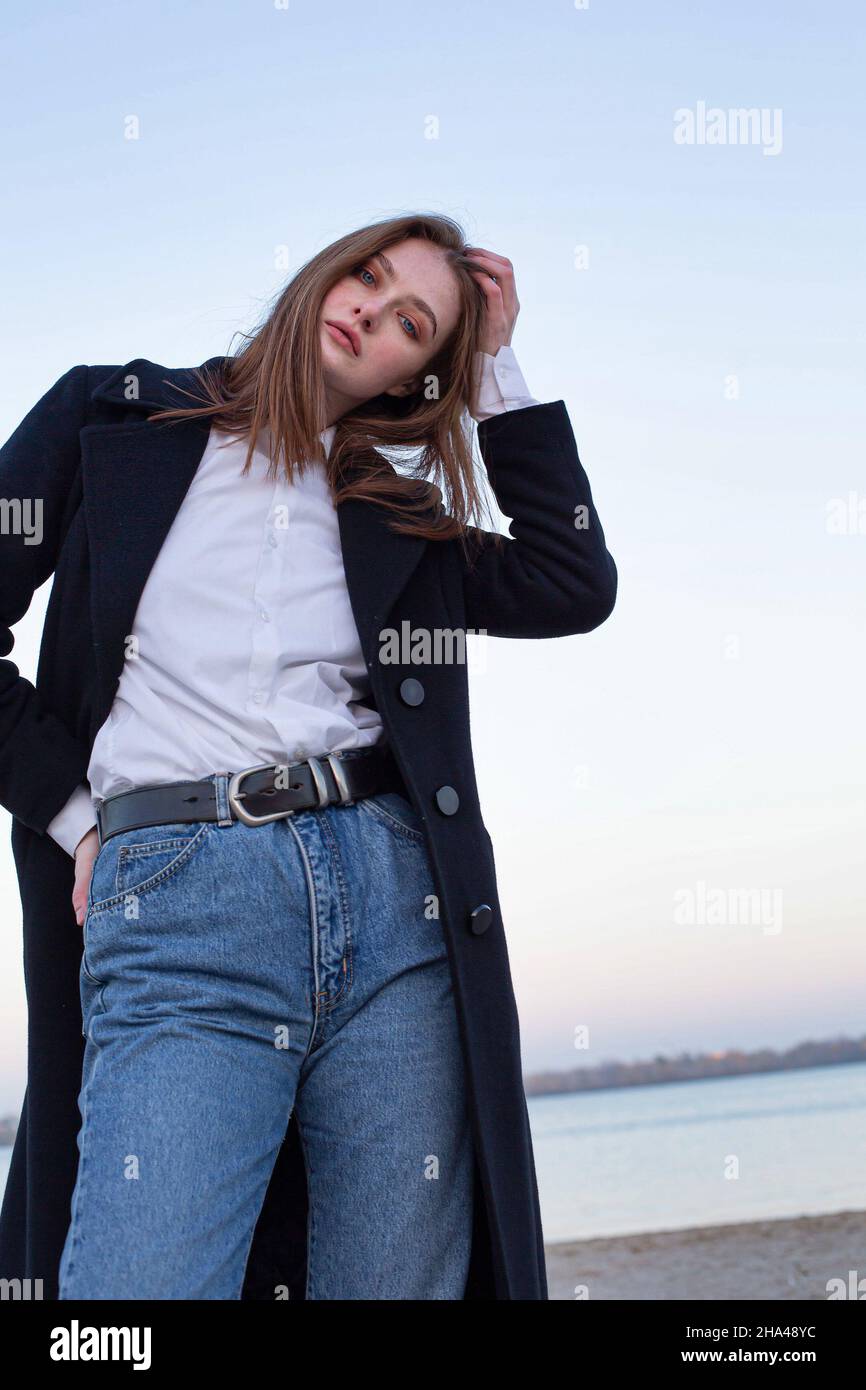 Long straight haired white european woman touching her head in dark coat in autumn on the beach Stock Photo
