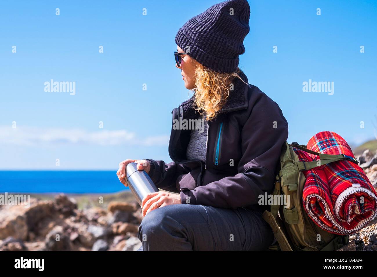 active adult lady resting and taking a break while do trekking adventure activity inthe nature. woman sit down on the rocks admiring ocean blue view. people travel with backpack Stock Photo