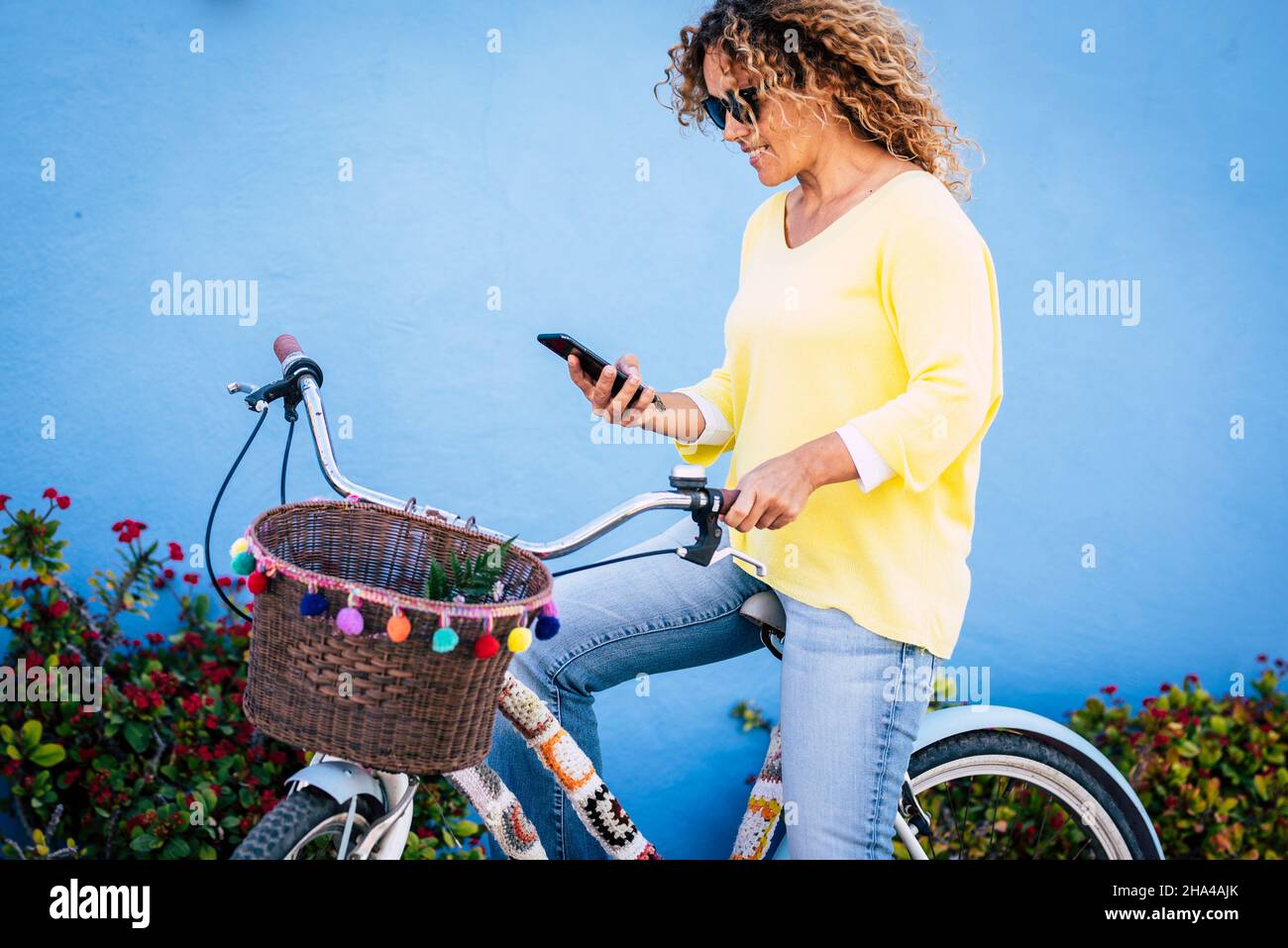 colorful yellow and blue image of young woman using cell phone sitting on a cute bike and blue wall in background with flowers. concept of happy leisure activity outdoor people Stock Photo