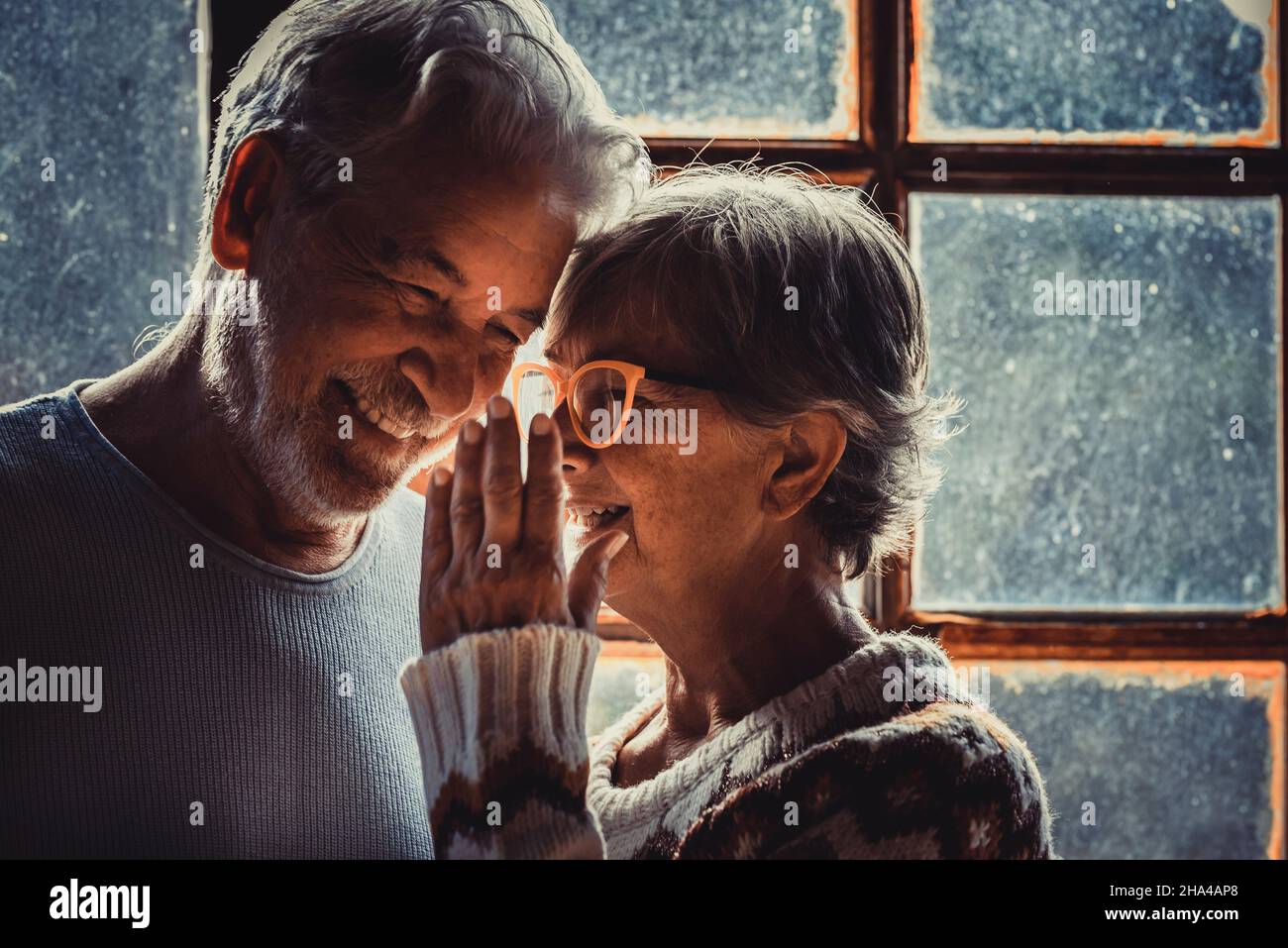 senior old couple in love at home with snow winter outside the window. aged woman whisper at man ear. mature couple enjoy relationship and holidays indoor. man and woman elderly smile Stock Photo