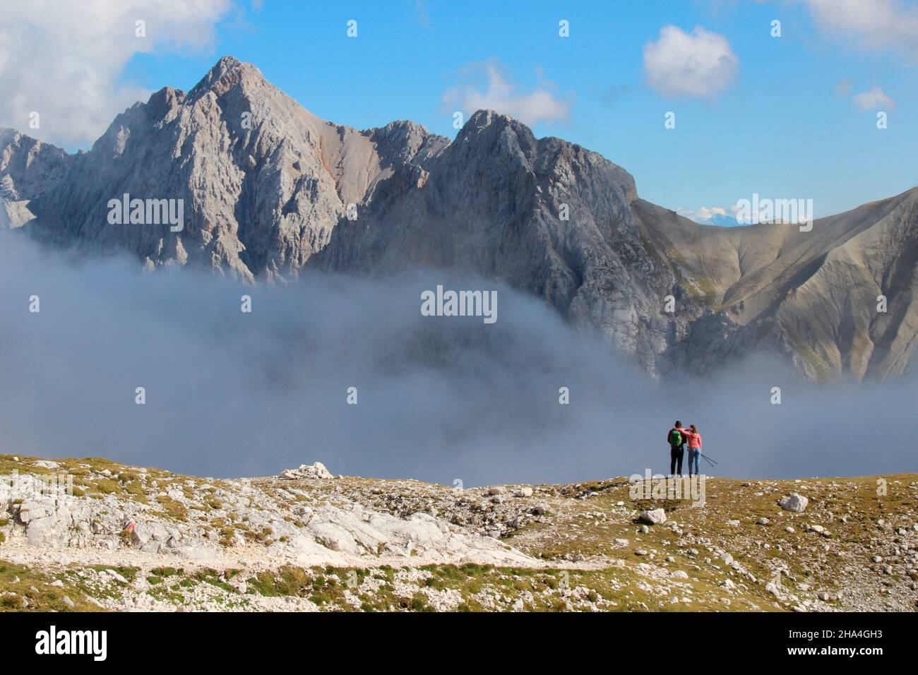 young couple hiking in the direction of zugspitze 2962 m,break with a fantastic view of the summit of hochwanner 2744m,wetterstein mountains blue sky,clouds,cloud mood,garmisch-partenkirchen,loisachtal,upper bavaria,bavaria,southern germany,germany,europe, Stock Photo