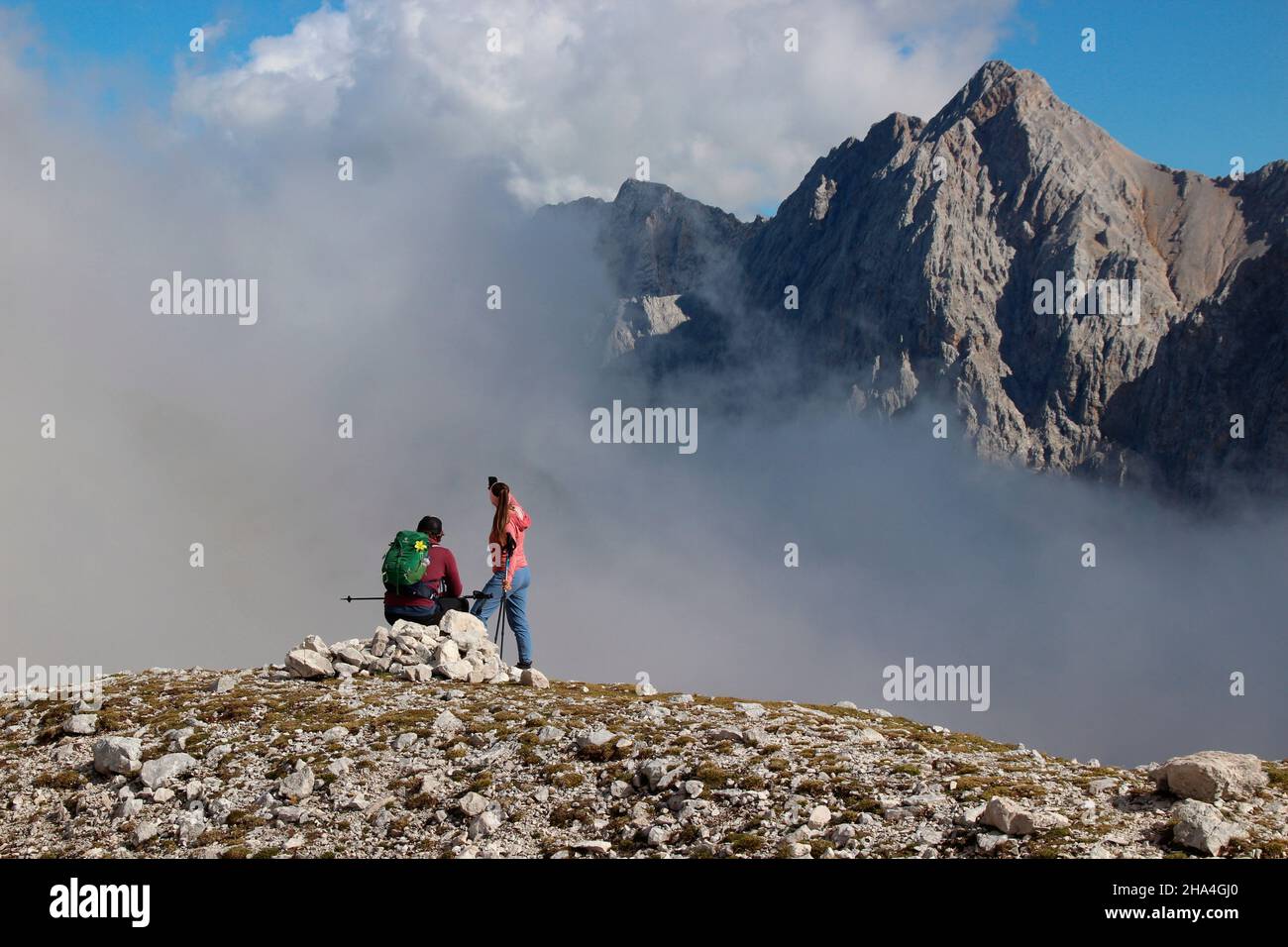 young couple hiking in the direction of zugspitze 2962 m,break with a fantastic view of the summit of hochwanner 2744m,wetterstein mountains blue sky,clouds,cloud mood,garmisch-partenkirchen,loisachtal,upper bavaria,bavaria,southern germany,germany,europe, Stock Photo