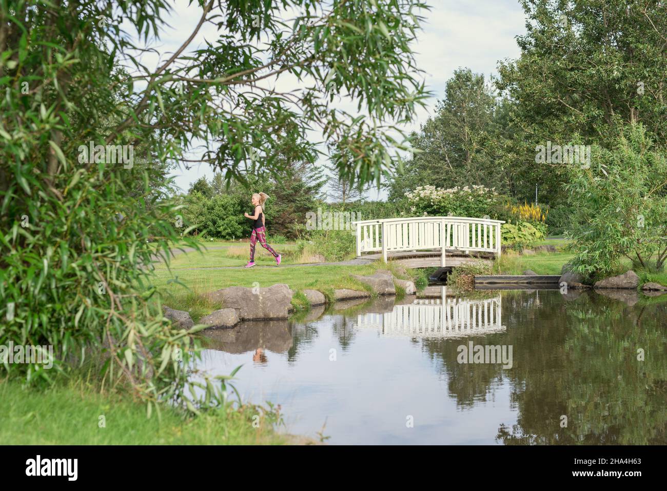 Side view of anonymous sportswoman with long blond hair in activewear running in green park near small bond and footbridge during outdoor training Stock Photo