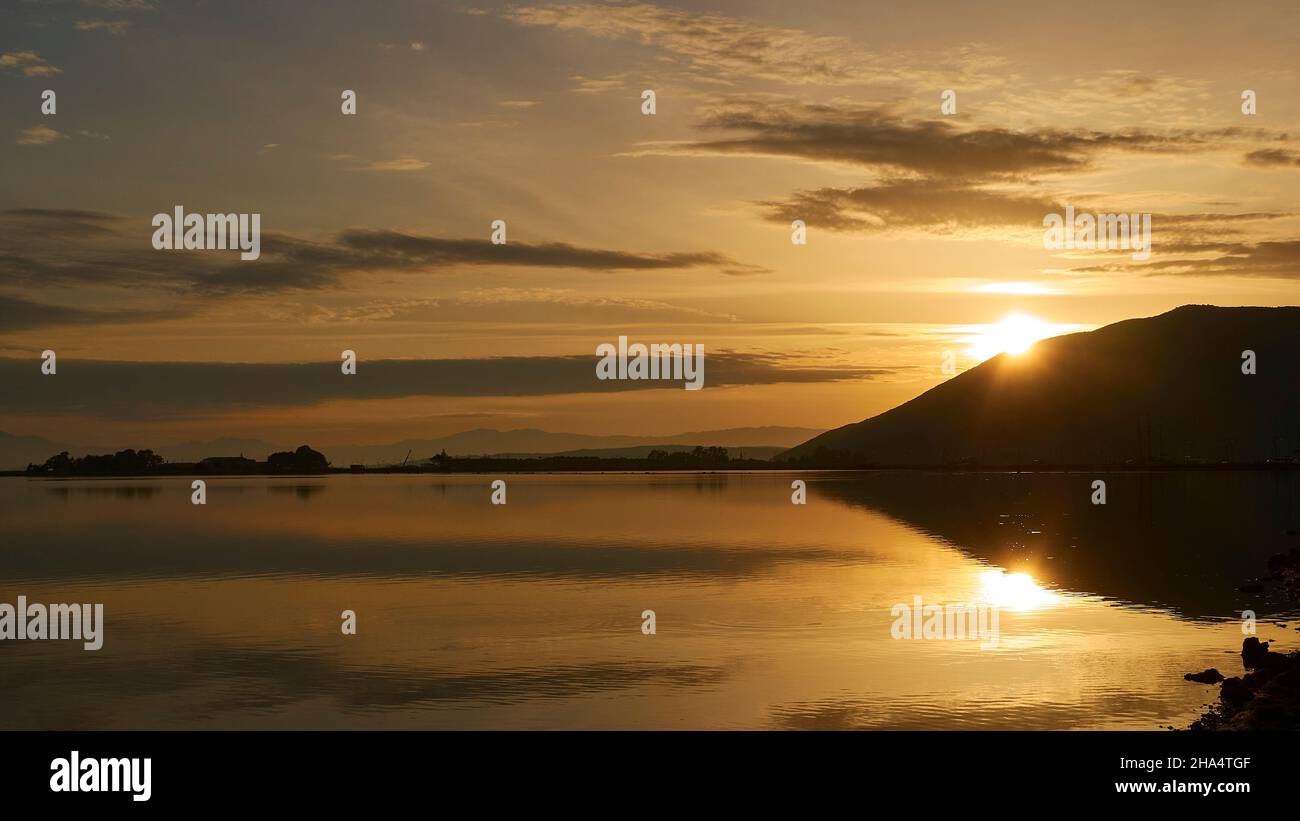 greece,greek islands,ionian islands,lefkada or lefkas,lefkada city,capital,morning light,dawn,sunrise,sun behind hill,sunbeams,sky light blue and orange with gray clouds,are reflected in the water Stock Photo