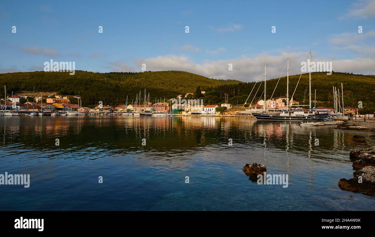 greece,greek islands,ionian islands,kefalonia,fiskardo,morning mood,partly cloudy sky,large part of the harbor basin,foreground in the shadow,boats in the middle distance,fiskardos houses in the morning light behind,gentle green chain of hills in the background,above blue sky with some gray-white clouds Stock Photo