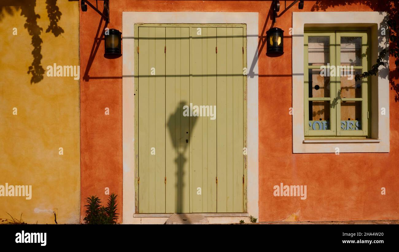 greece,greek islands,ionian islands,kefalonia,fiskardo,morning mood,house front directly on the promenade,detail,facade is ocher and red,door and windows are green,shadow of a street lamp on the green door Stock Photo