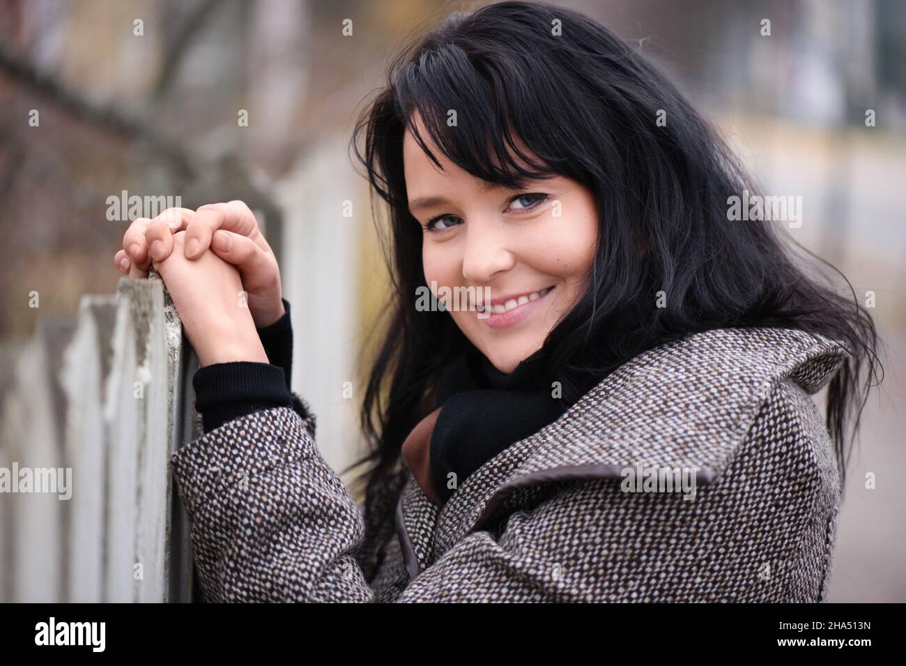 Smiling young woman in jacket standing beside old picket fence. Selective focus and shallow depth of field. Stock Photo