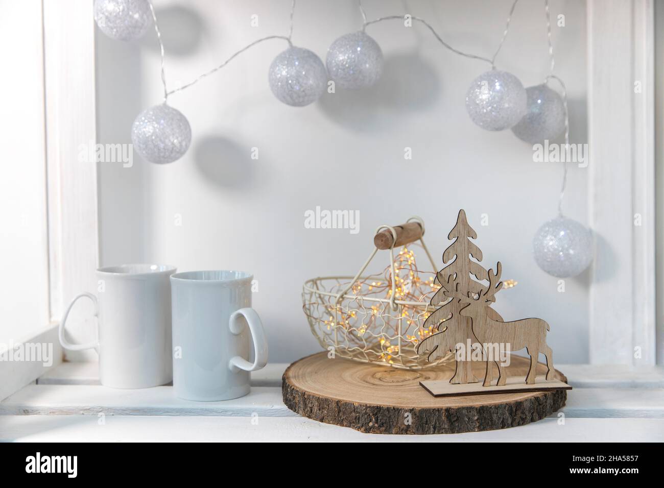 A wooden figurine of deer and spruce against background of small chest of drawers, a metal decorative basket, two mugs of tea, a silver garland of thr Stock Photo
