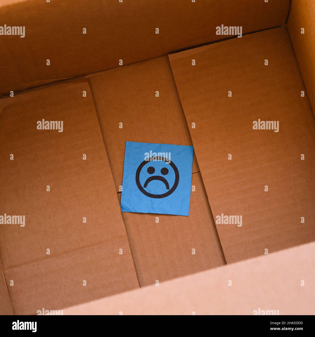 A blue paper note with a sad face on it in a cardboard box. Close up. Stock Photo