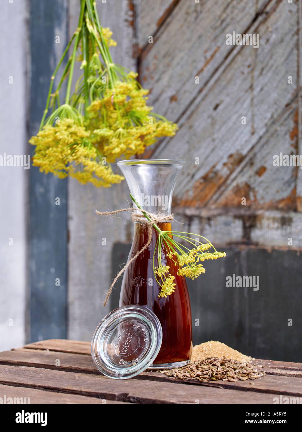 work steps with fennel and fennel seeds for the preparation of fennel syrup,wake-up bottle with fennel syrup on a wooden table in front of an old wooden door on which the tied branches of fennel hang Stock Photo