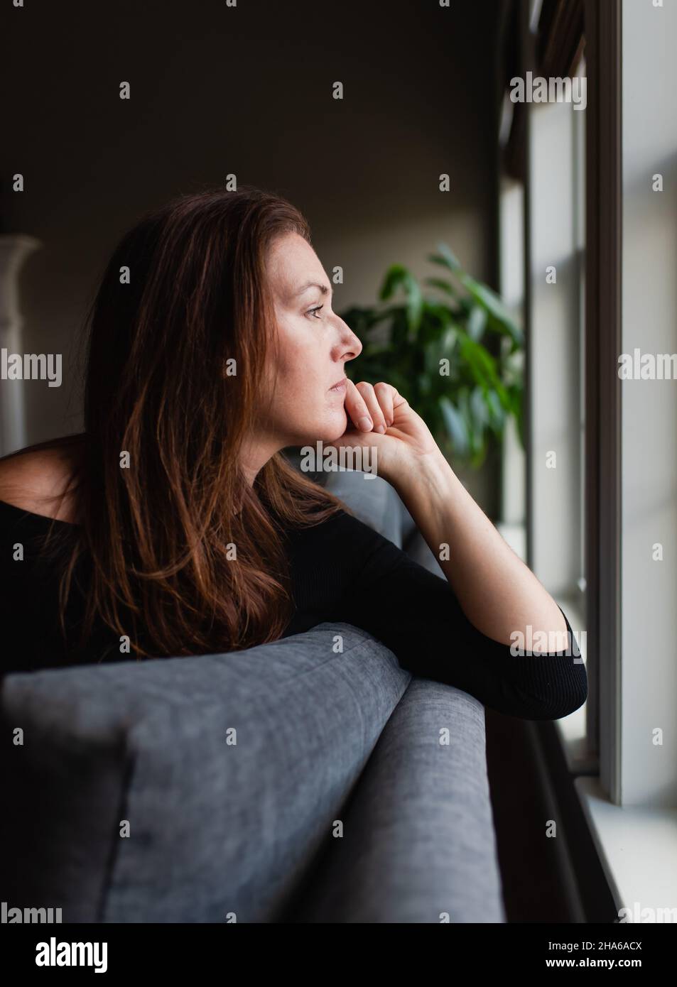 Woman looking thoughtfully out window while sitting on sofa. Stock Photo