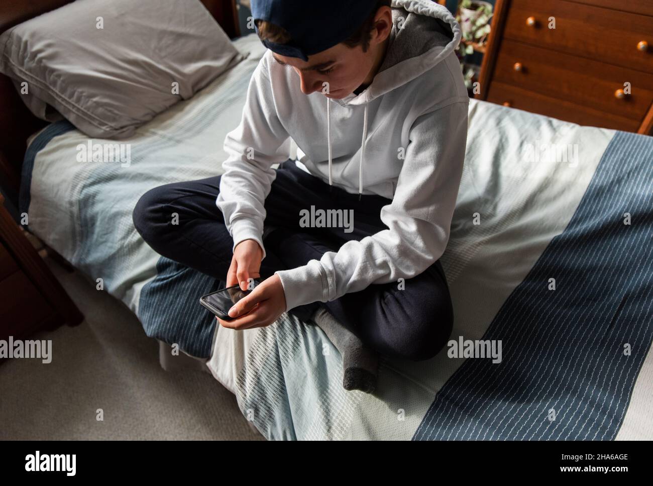High angle of teen boy in white sweater on smartphone in bedroom. Stock Photo