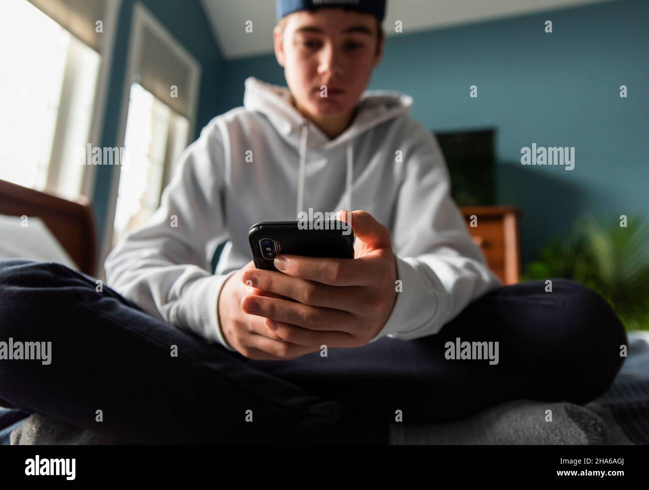 Low angle of teen boy in white sweater on smartphone in bedroom. Stock Photo