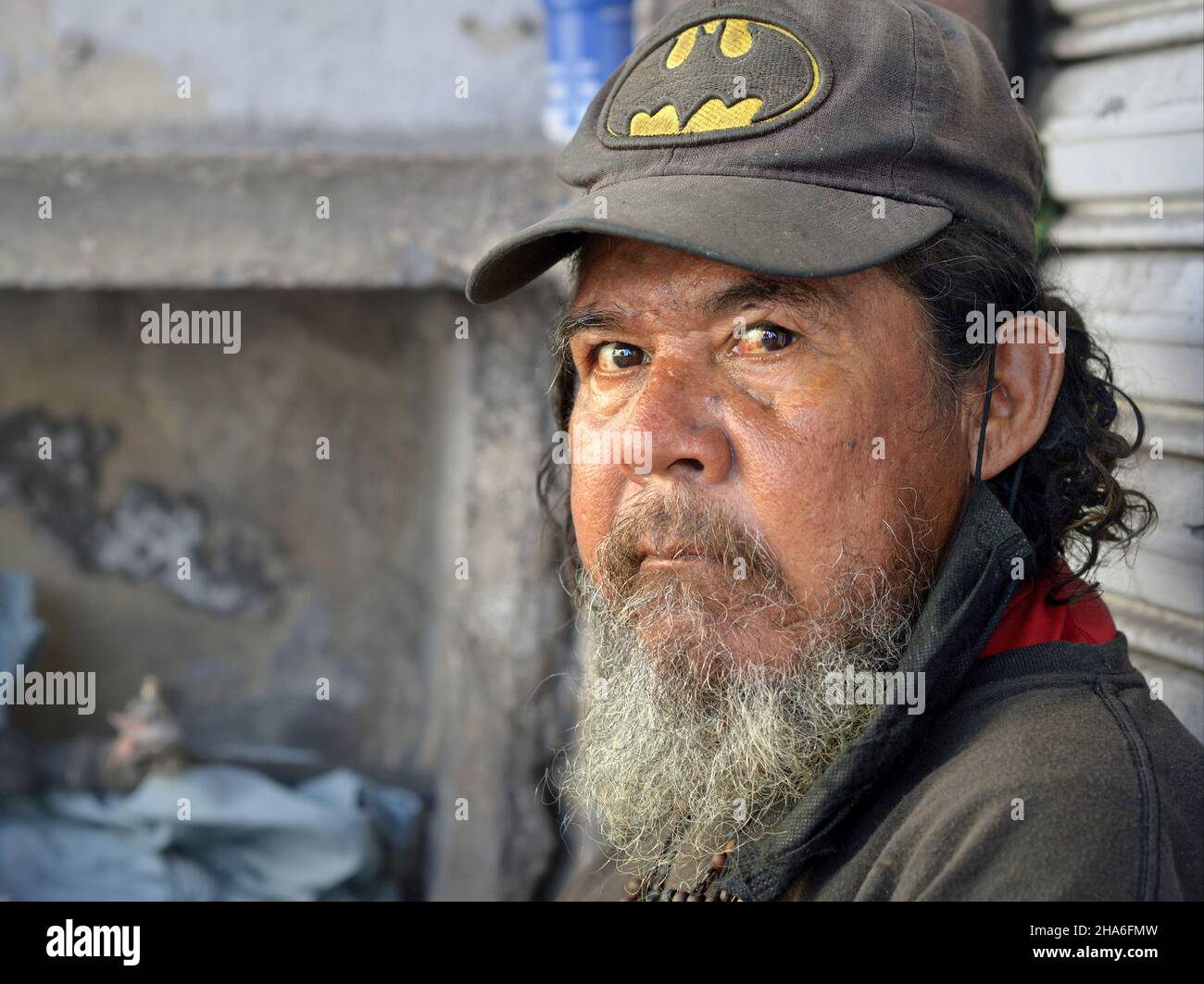 Elderly rugged homeless Mexican man with grey beard and disheveled hair wears a worn baseball cap and looks at the viewer. Stock Photo