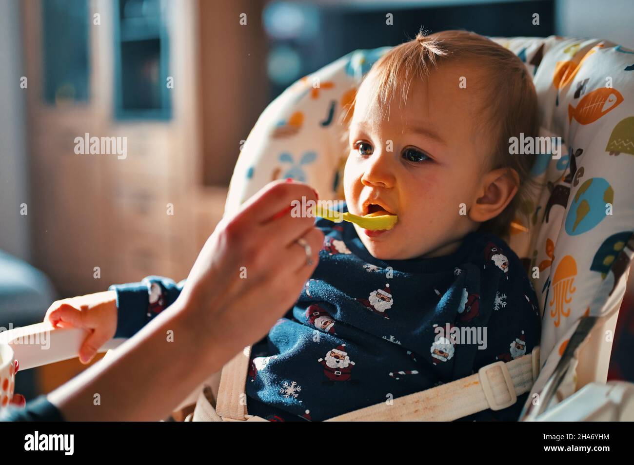 Adorable little baby sitting in high chair while his mother feeding him Stock Photo