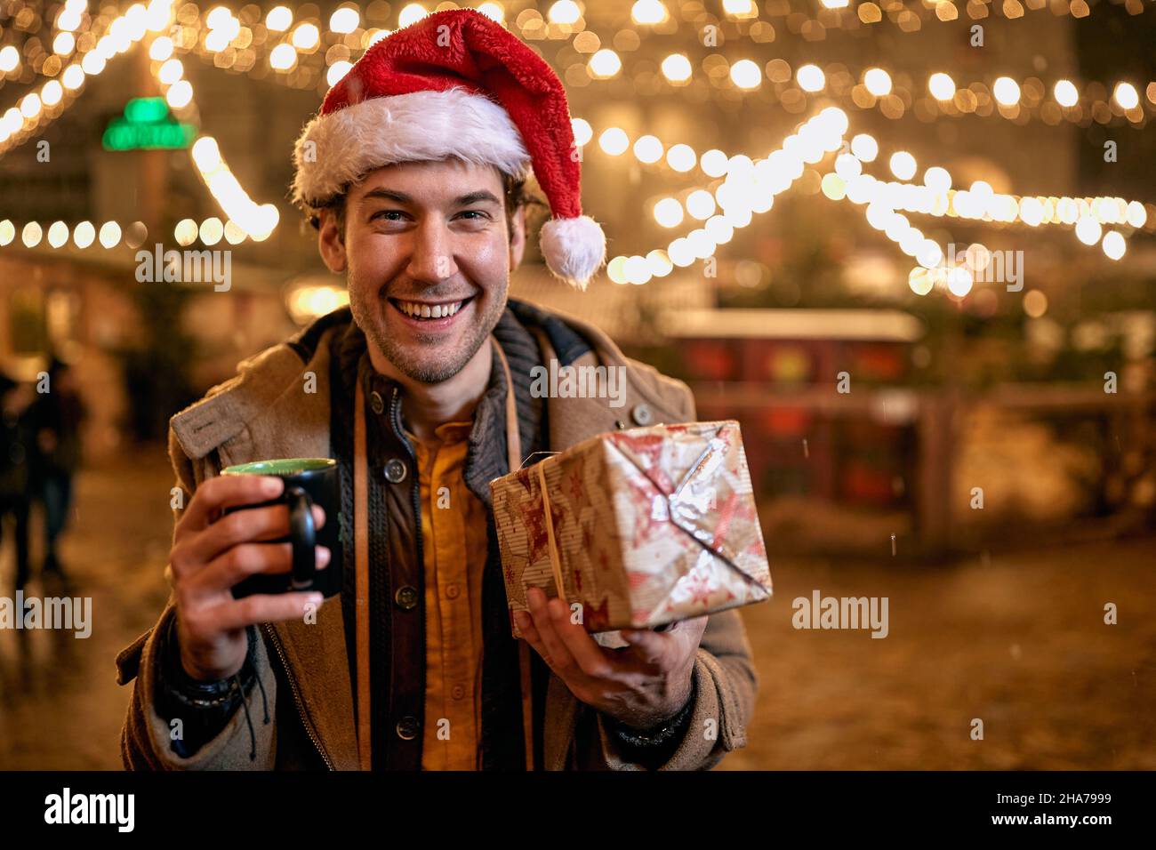 Smiling young man in santa hat drinking tea outdoors at winter and giving Christmas gift. Stock Photo