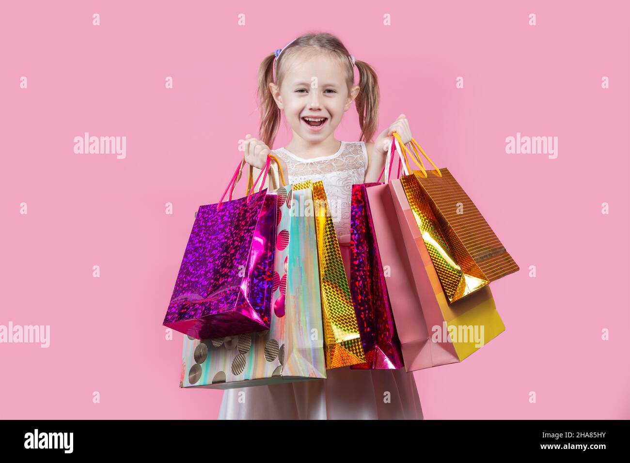 Funny little caucasian girl in a party dress with shopping bags isolated over pink background with copy space. Stock Photo
