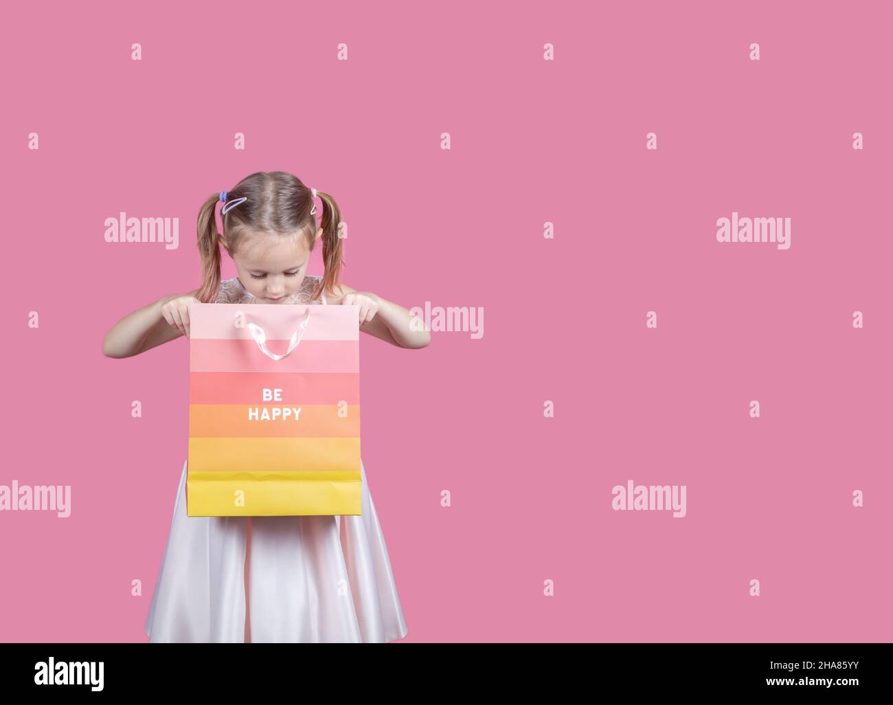 Portrait of an excited little girl wearing dress and holding colorful shopping bags isolated over pink background with copy space. Stock Photo
