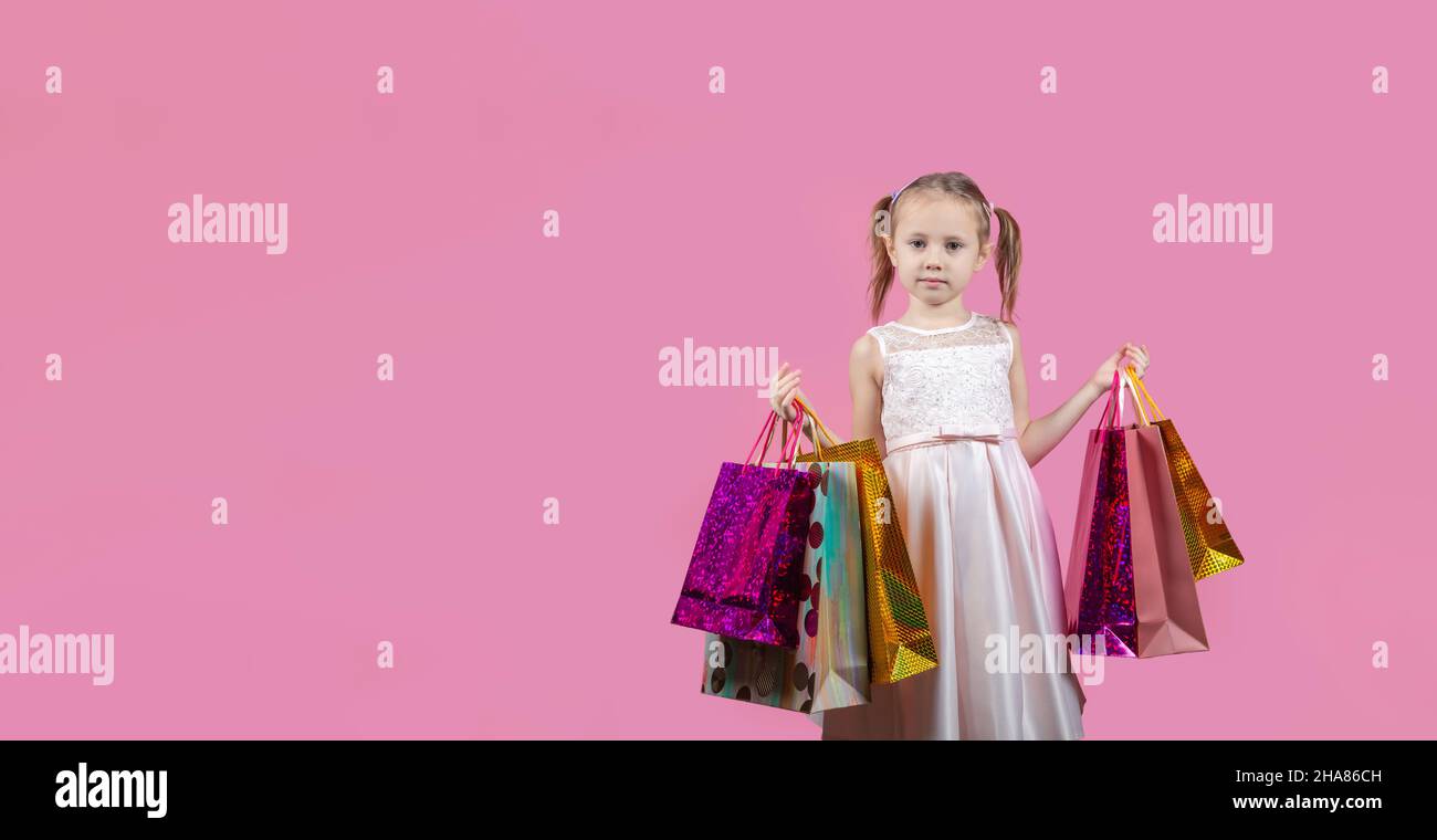 Cute little caucasian girl in a party dress on pink background happy enjoying shopping holding colorful bags Stock Photo