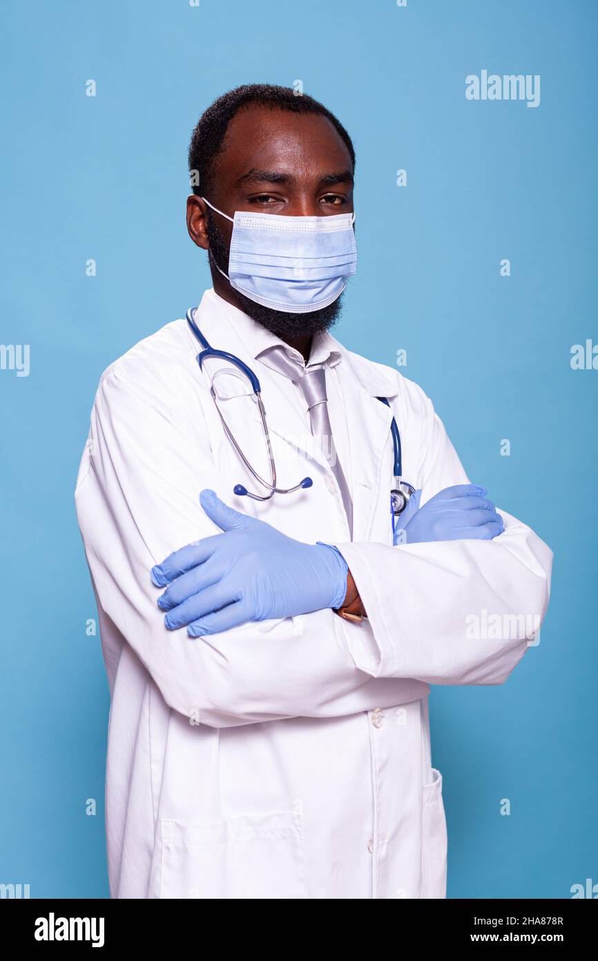 Vertical portrait of confident medic with arms crossed wearing face mask and latex gloves before scrubbing in. Doctor with stethoscope wearing protective medical gear and white lab coat. Stock Photo