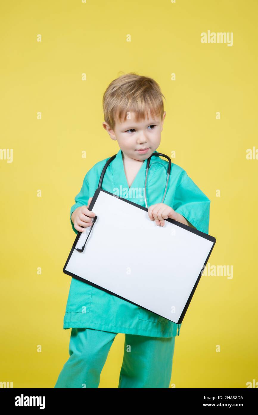 Portrait of little caucasian kid dressed in doctors green coat holds tablet in hand isolated on yellow Stock Photo
