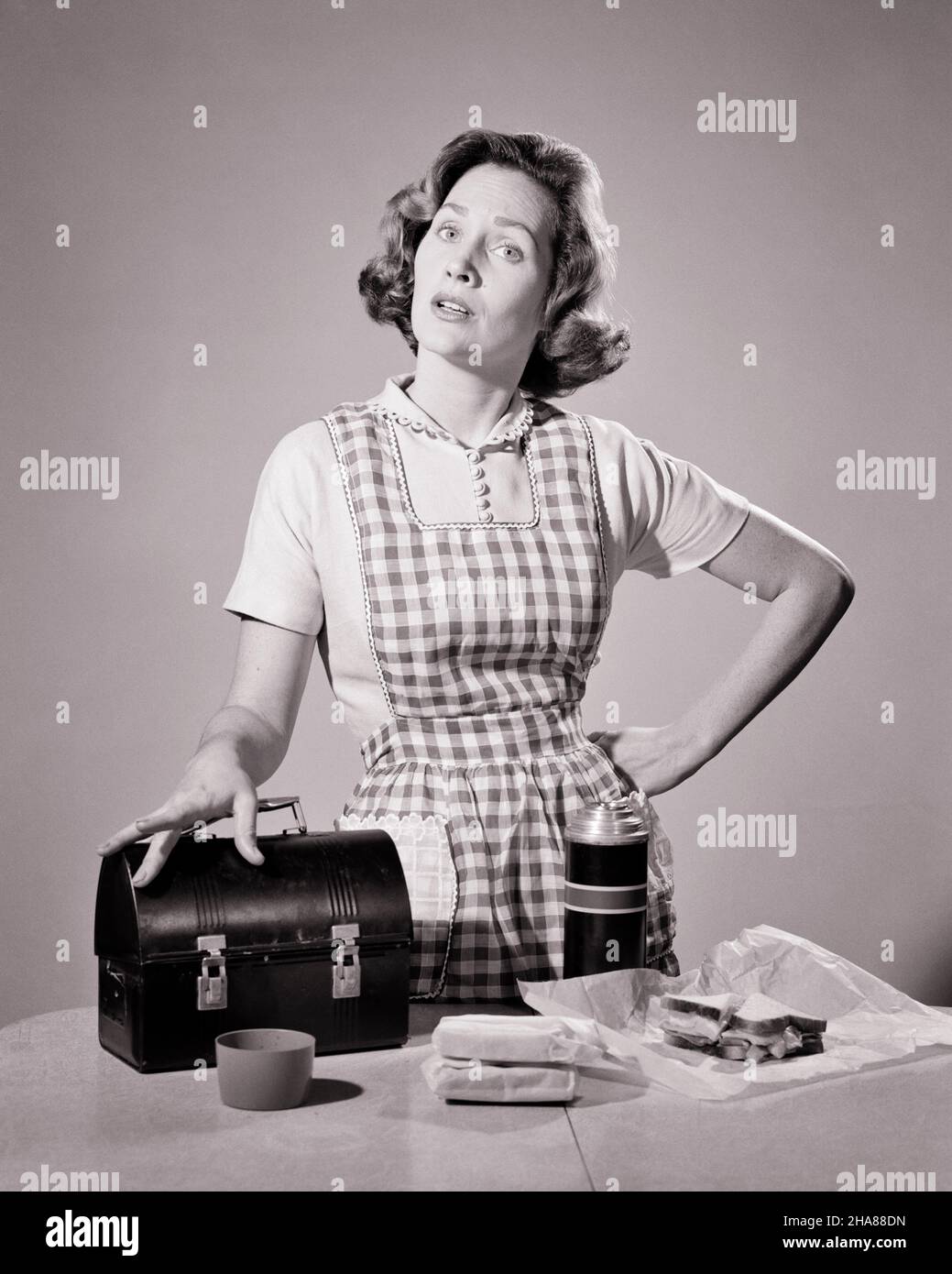 1950s 1960s SERIOUS WOMAN HOUSEWIFE LOOKING AT CAMERA WEARING CHECKERED APRON PREPARING MEAL TO PACK IN LUNCH BOX AND THERMOS - h3319 DEB001 HARS 1 FACIAL YOUNG ADULT WORRY LIFESTYLE FEMALES MOODY PACK HOME LIFE PREPARING HALF-LENGTH LADIES PERSONS CARING CONFIDENCE EXPRESSIONS TROUBLED B&W CONCERNED SADNESS EYE CONTACT HOMEMAKER HOMEMAKERS PAIL AND THERMOS PRIDE IN TO HOUSEWIVES MOOD CONCEPTUAL GLUM STYLISH SUPPORT DEB001 CHECKERED MISERABLE YOUNG ADULT WOMAN BLACK AND WHITE CAUCASIAN ETHNICITY OLD FASHIONED Stock Photo